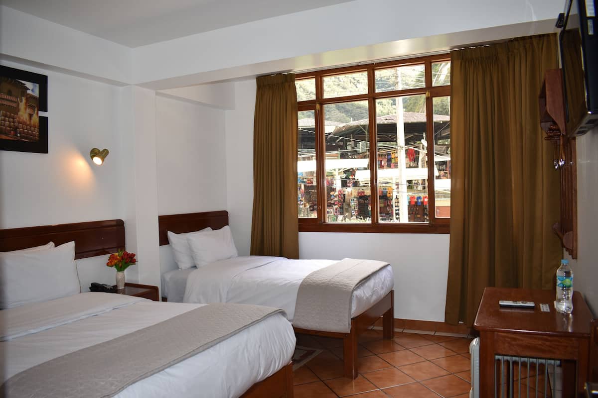 A hotel room with two beds, a wooden nightstand with a vase of flowers, a wooden table with a bottle, and a window with curtains.