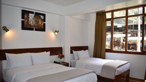 A hotel room with two beds, a nightstand with a phone and vase, a wall-mounted picture, and a window with a view of the outside.