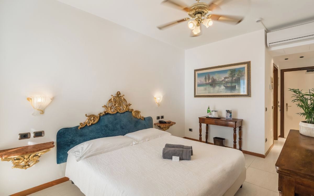 A hotel room with a large bed, a ceiling fan, a painting on the wall, and a small table with a plant.