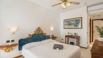 A hotel room with a large bed, a ceiling fan, a painting on the wall, and a small table with a plant.