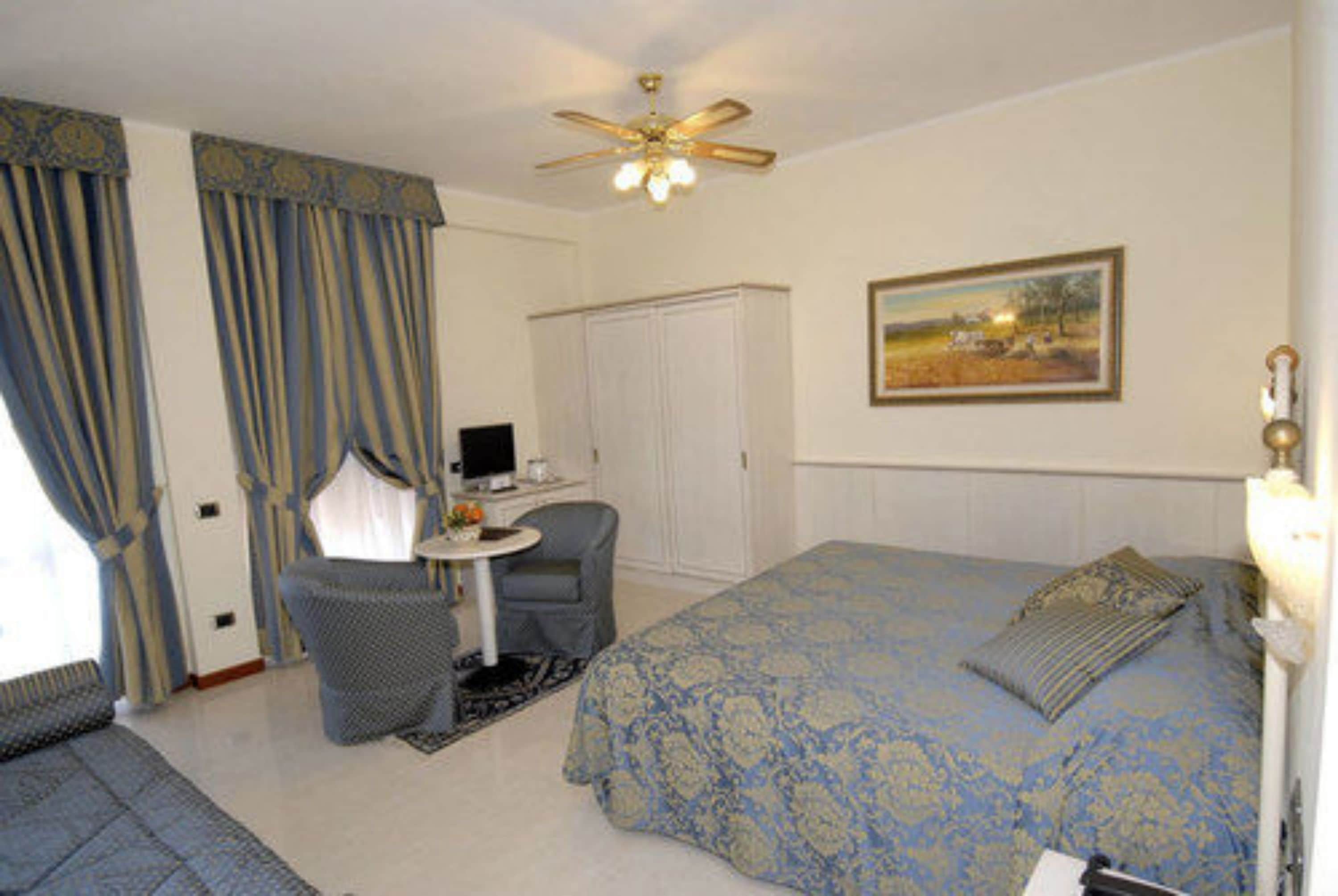 Quarto de hotel com cama, escrivaninha, cadeira, ventoinha no teto e um quadro na parede.