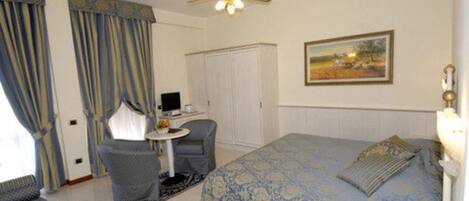 Quarto de hotel com cama, escrivaninha, cadeira, ventoinha no teto e um quadro na parede.