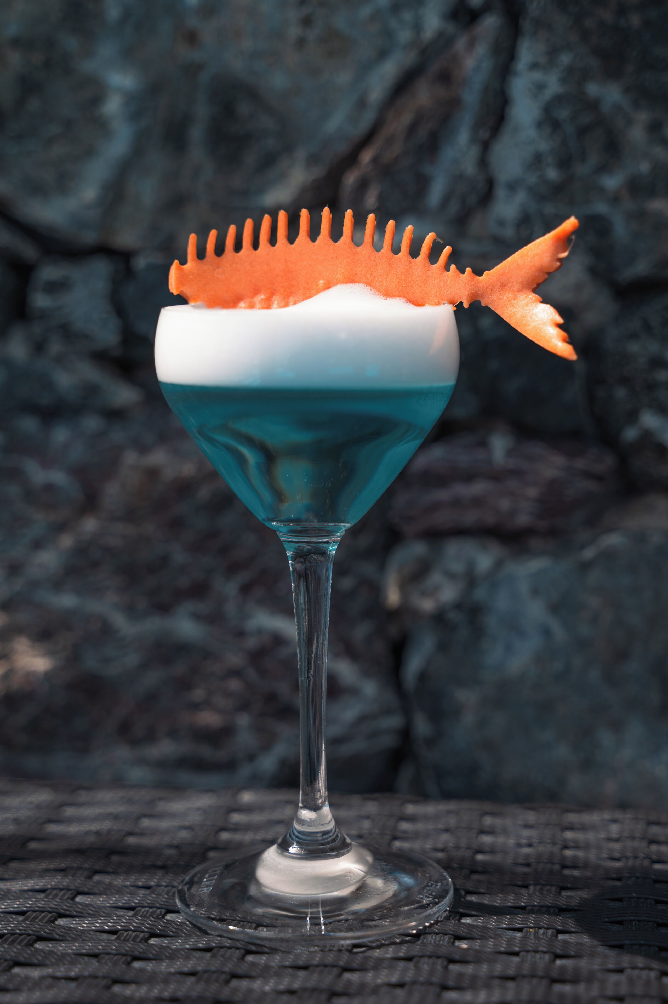 Un cocktail servi dans un verre à martini, agrémenté d’une garniture d’orange originale, le tout présenté sur un fond de pierre à texture marquée.