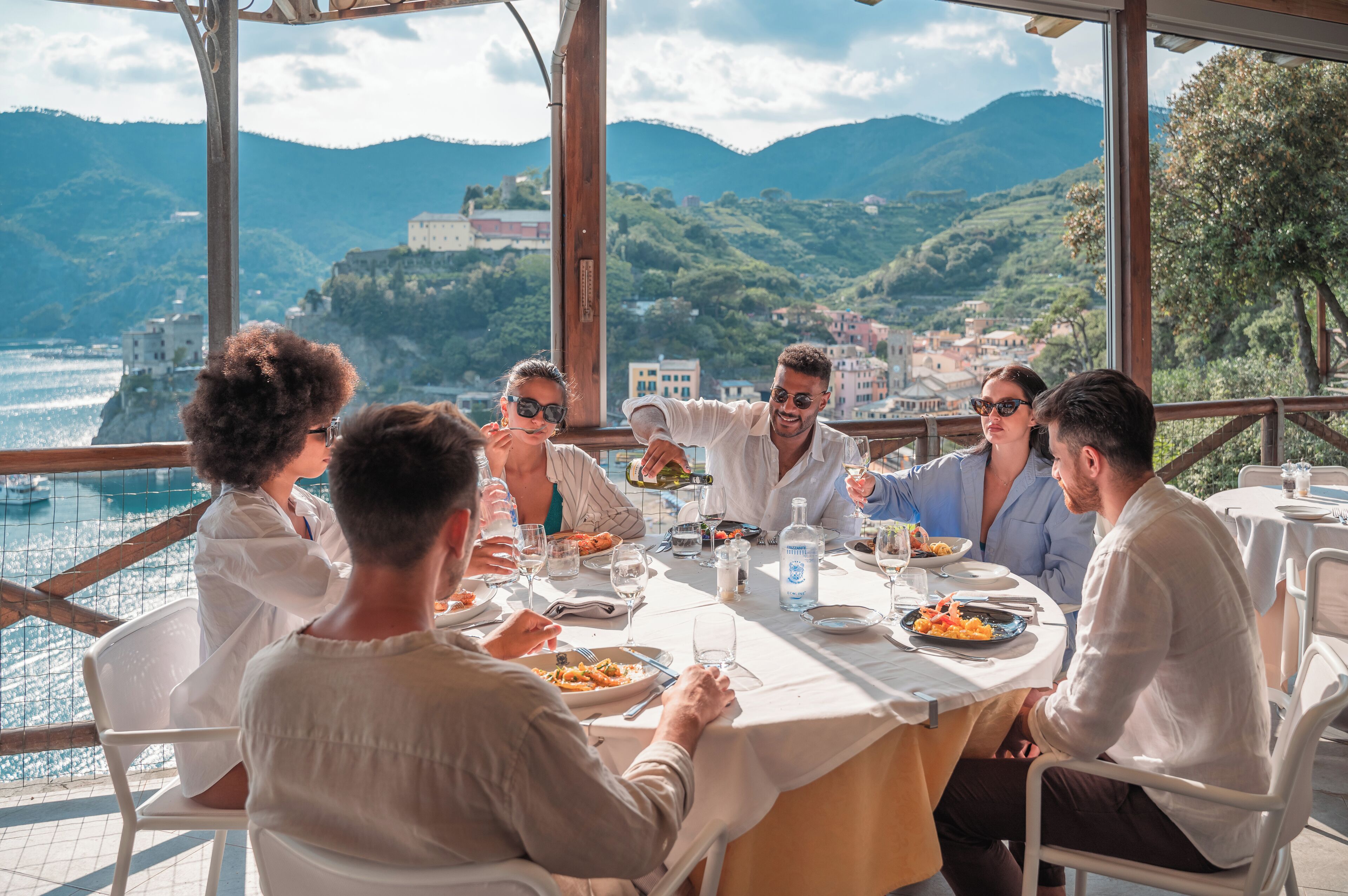 Un gruppo di persone cena a un tavolo con vista sul mare e sulle montagne.