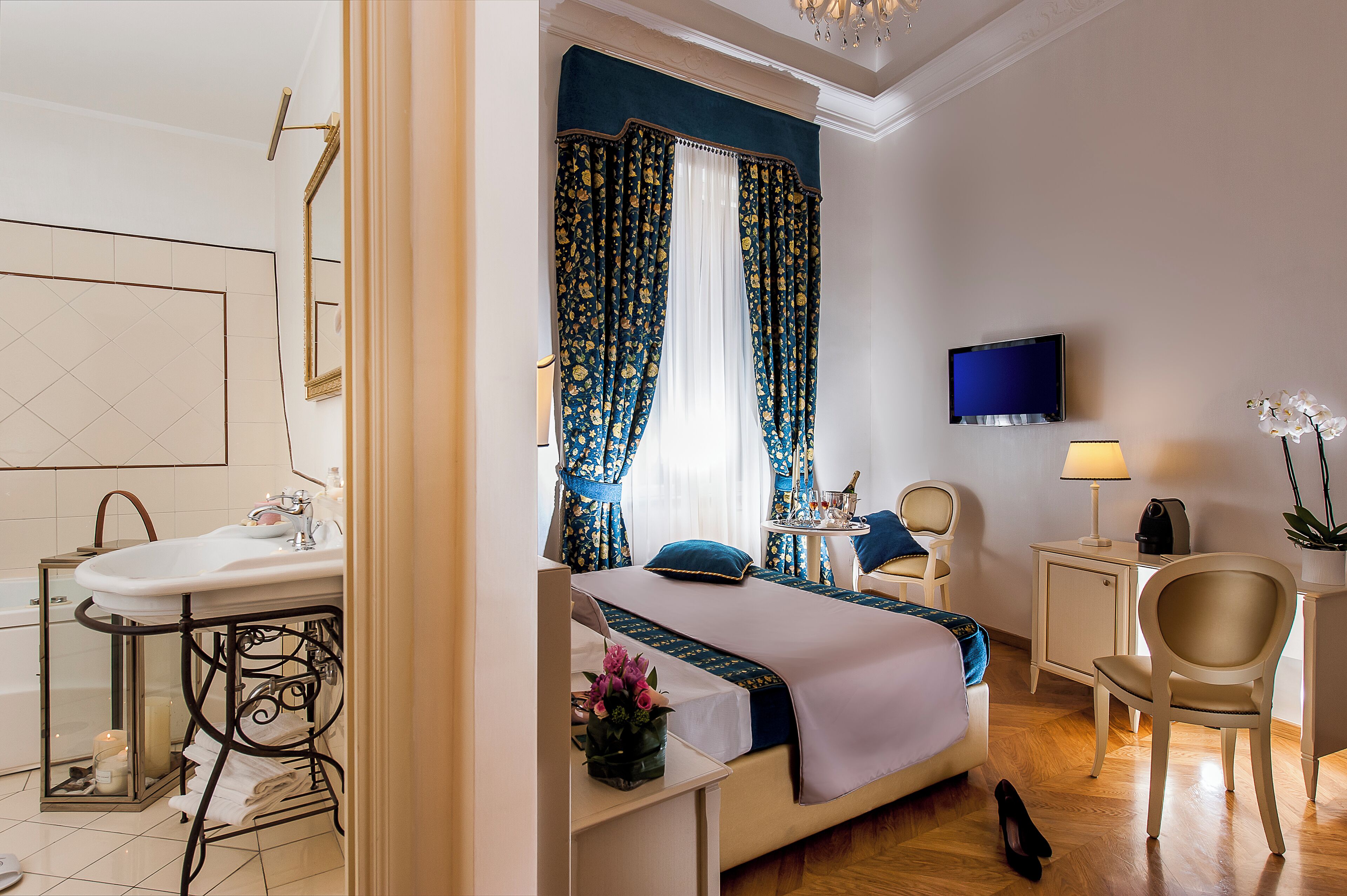 Habitación de hotel con cama, escritorio, silla, televisor, lámpara y baño con lavamanos y espejo.