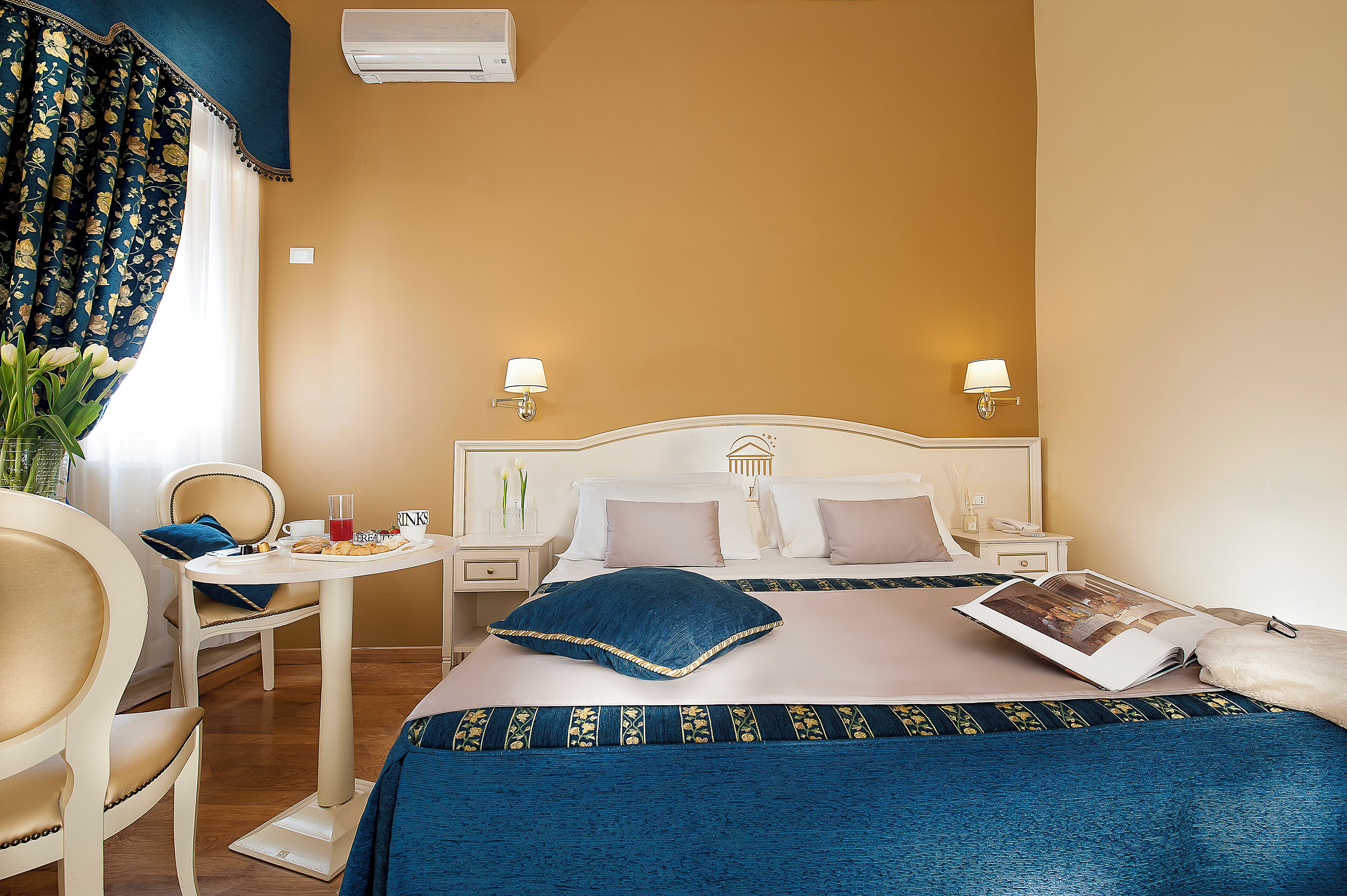 A hotel room with a bed, bedside tables, a chair, a small table, and a window with curtains.