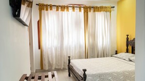 Quarto com cama, cortinas, um banco de madeira e uma TV fixada na parede.