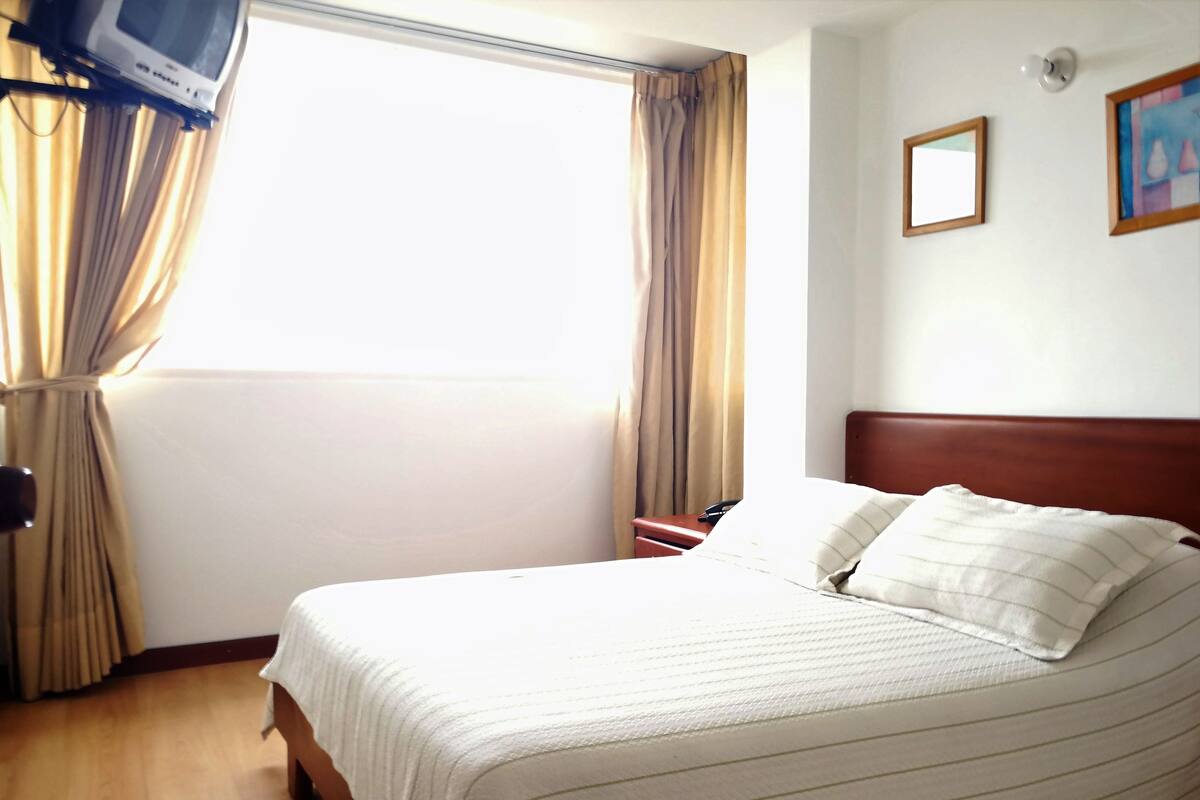 A hotel room with a bed, curtains, a bedside table, a telephone, and a wall-mounted light fixture.