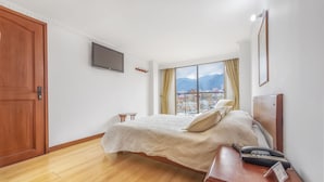 A hotel room with a wooden floor, a bed with a white bedspread, a mounted TV, a window with curtains, and a city view. - Hotel Alicante (Bogotá)