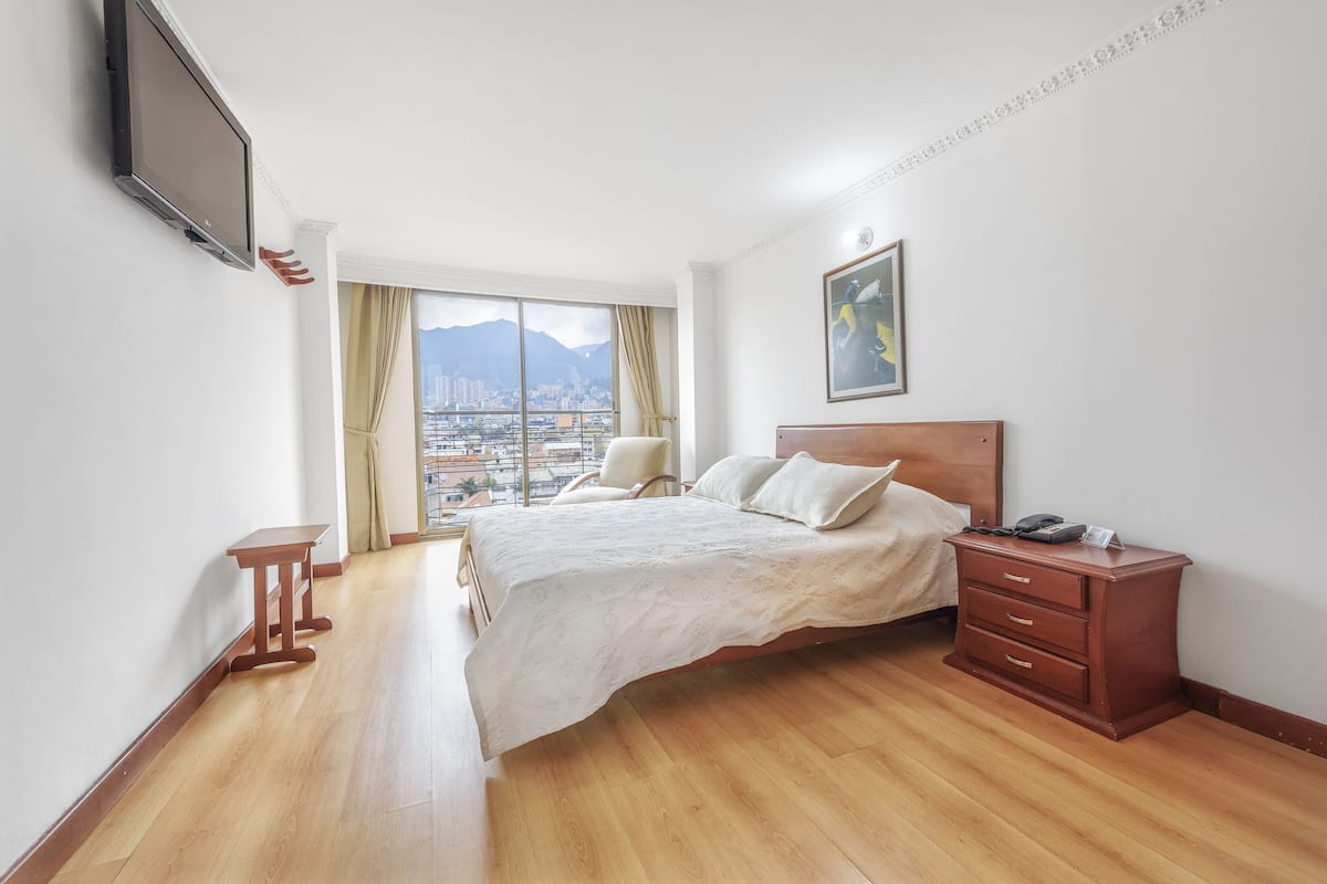 A hotel room with a bed, a bedside table, a lamp, a TV mounted on the wall, and a window with a city view.