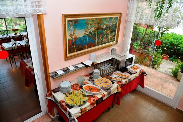 A buffet setup with various food items, a painting on the wall, and a view of the outdoors through a window. - Hostal Magnolia (Lloret de Mar)