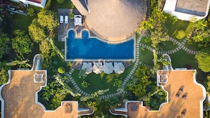 An aerial view of a resort with a central swimming pool, surrounded by lush greenery and a sandy beach area.