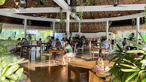 An indoor dining area with wooden tables and chairs, surrounded by hanging plants and a thatched roof ceiling.