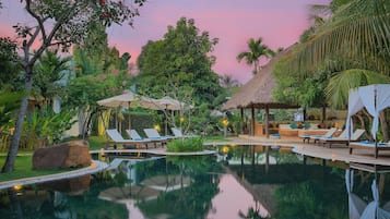 Un área de piscina tranquila con sillones de descanso, una estructura de techo de paja y abundante vegetación.
