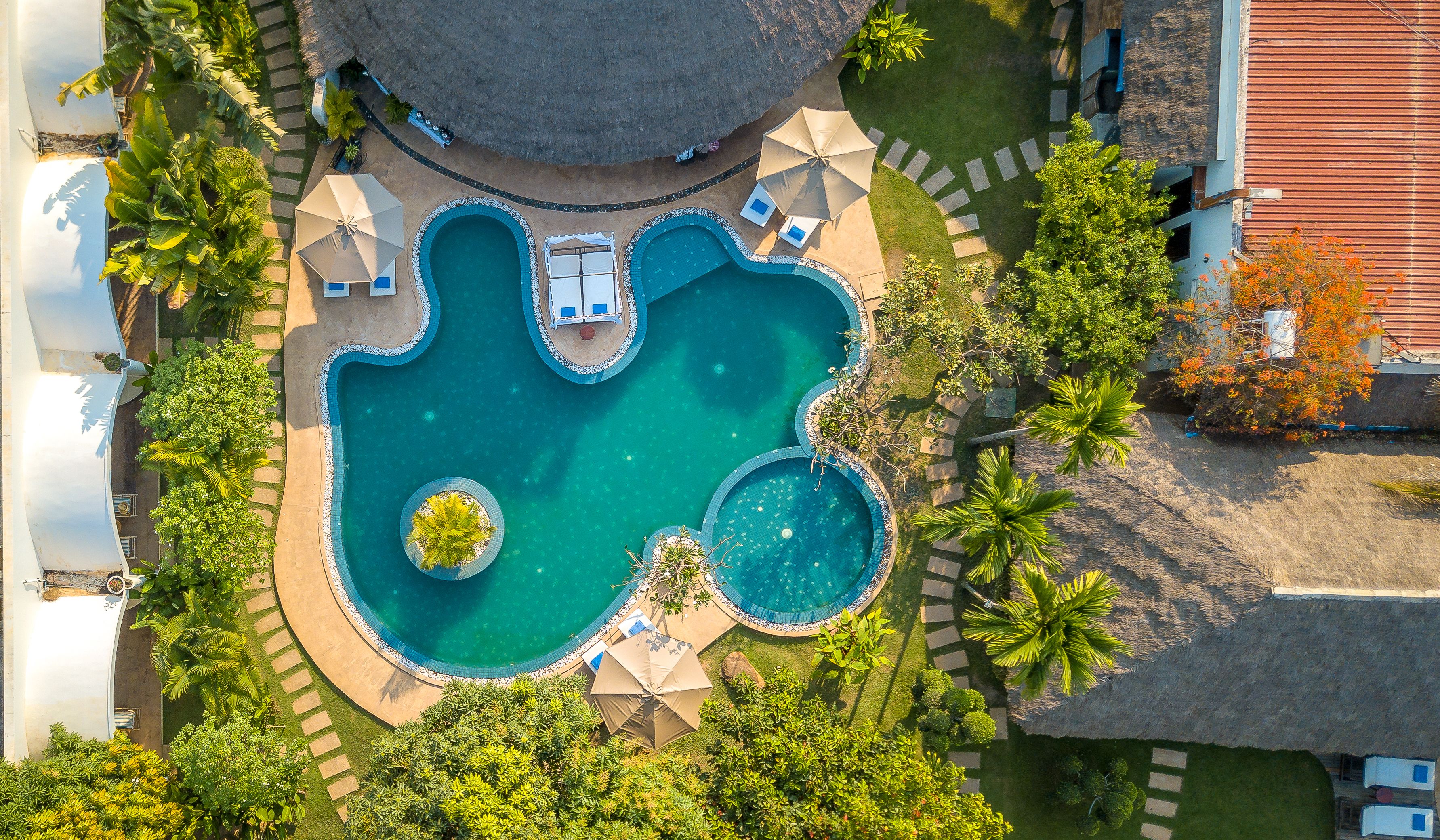 An aerial view of a resort with a central swimming pool, surrounded by lounge chairs and umbrellas, and lush greenery.