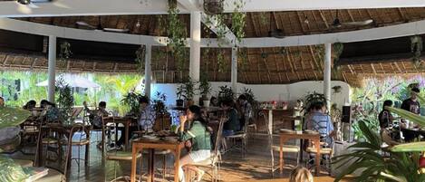 Un espace de restauration en plein air doté de toits de chaume, de tables et de chaises en bois, le tout entouré d’une végétation luxuriante.