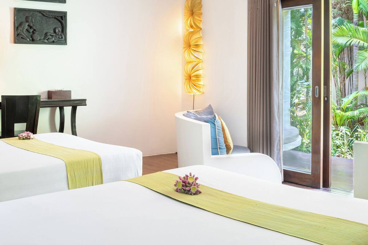 A hotel room with two beds, a desk, a chair, a lamp, and a view of a lush garden.