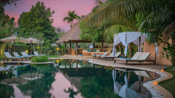A serene pool area with lounge chairs and a thatched-roof structure, surrounded by lush greenery.