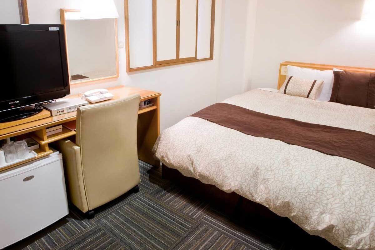 A hotel room with a bed, a desk with a television, a chair, a small refrigerator, and a bedside table.