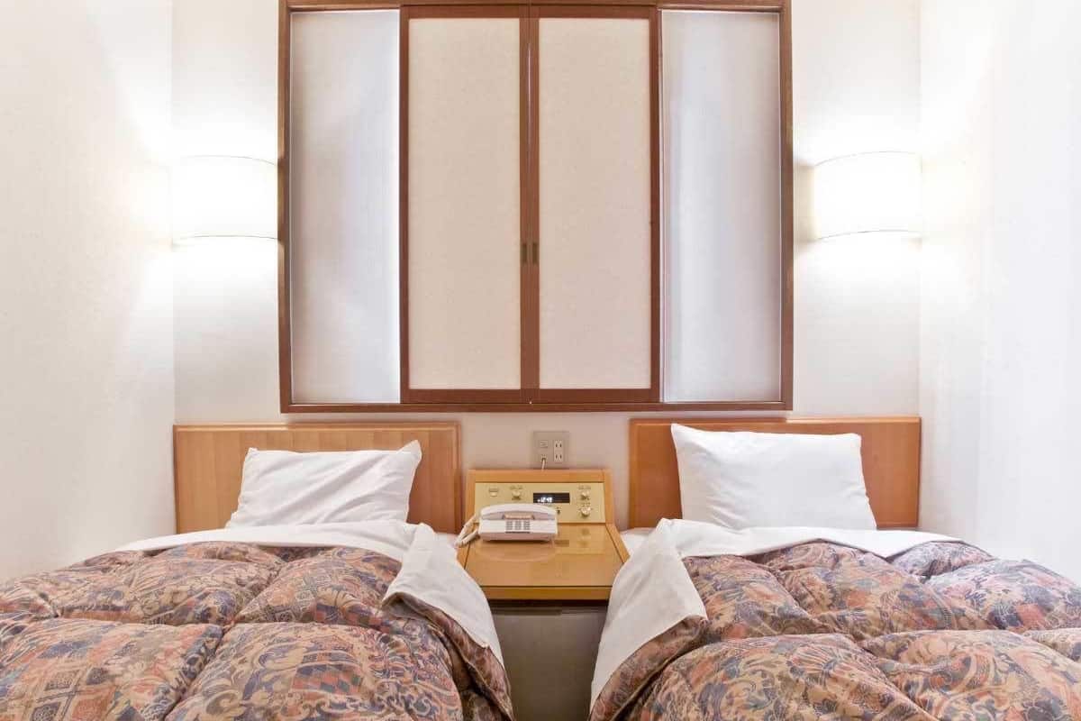 A hotel room with two beds, a wooden headboard, a bedside table with a clock, and a sliding door with frosted glass panels.