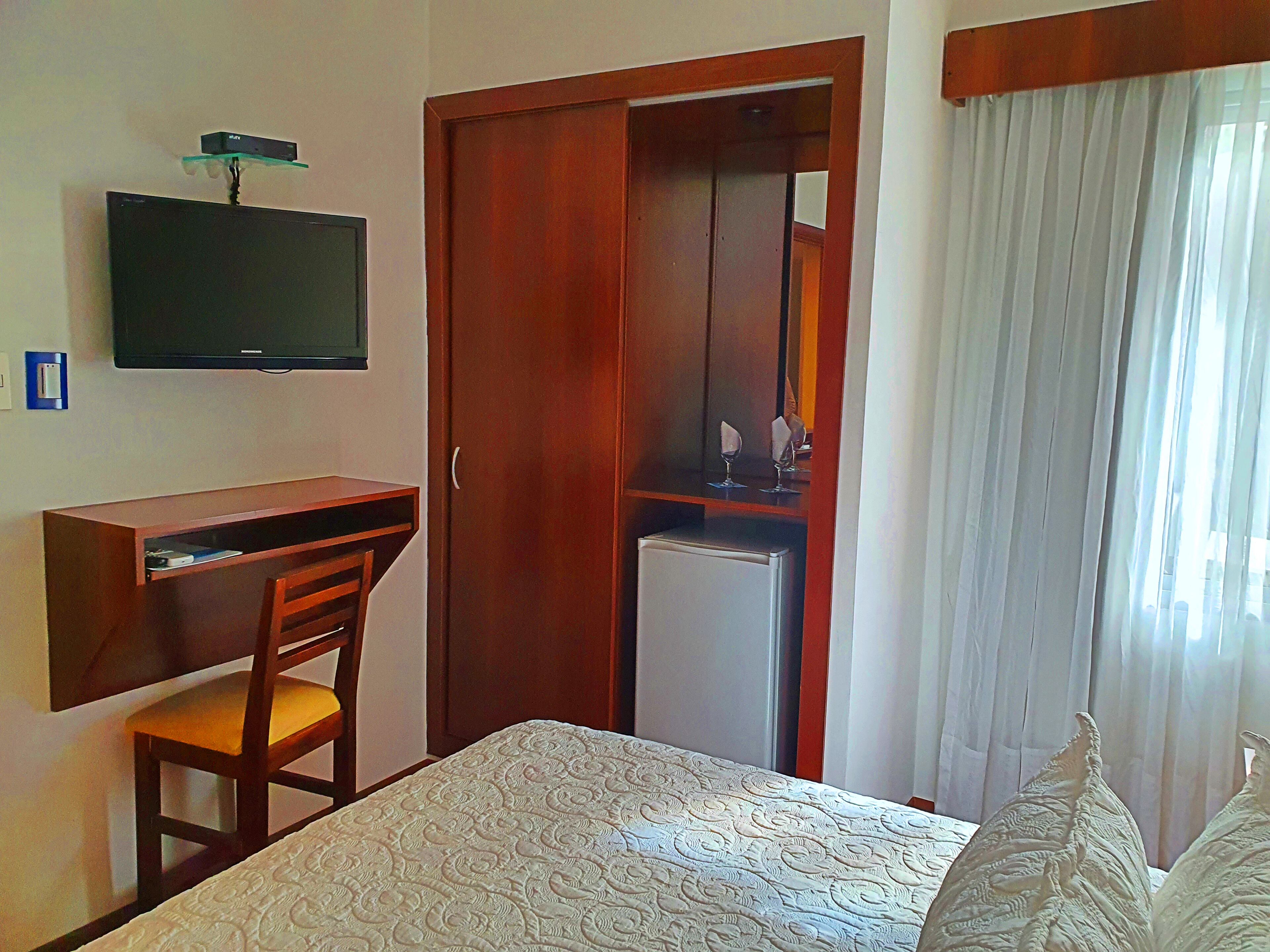 Habitación de hotel con cama, escritorio y silla, televisor montado en la pared y armario con espejo.