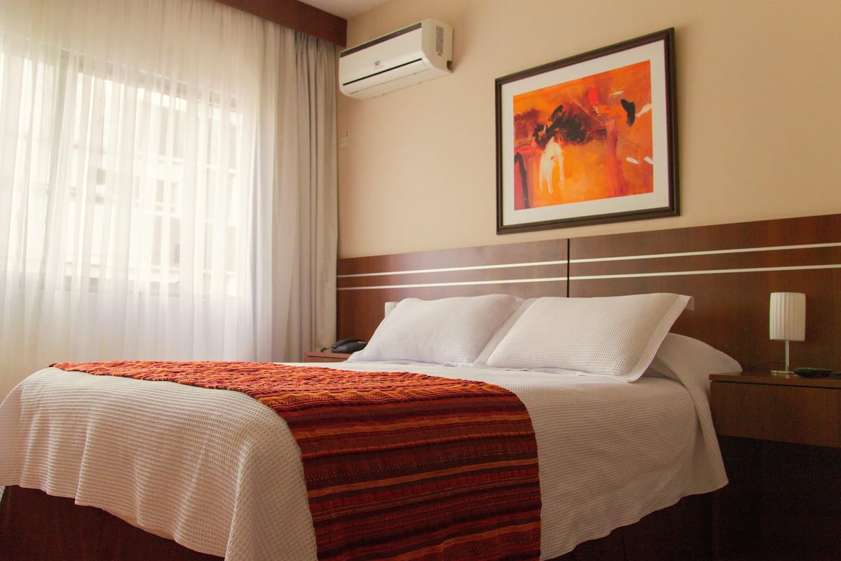 Una habitación de hotel con cama, mesita de noche, equipo de aire acondicionado, un cuadro en la pared y una ventana con cortinas.