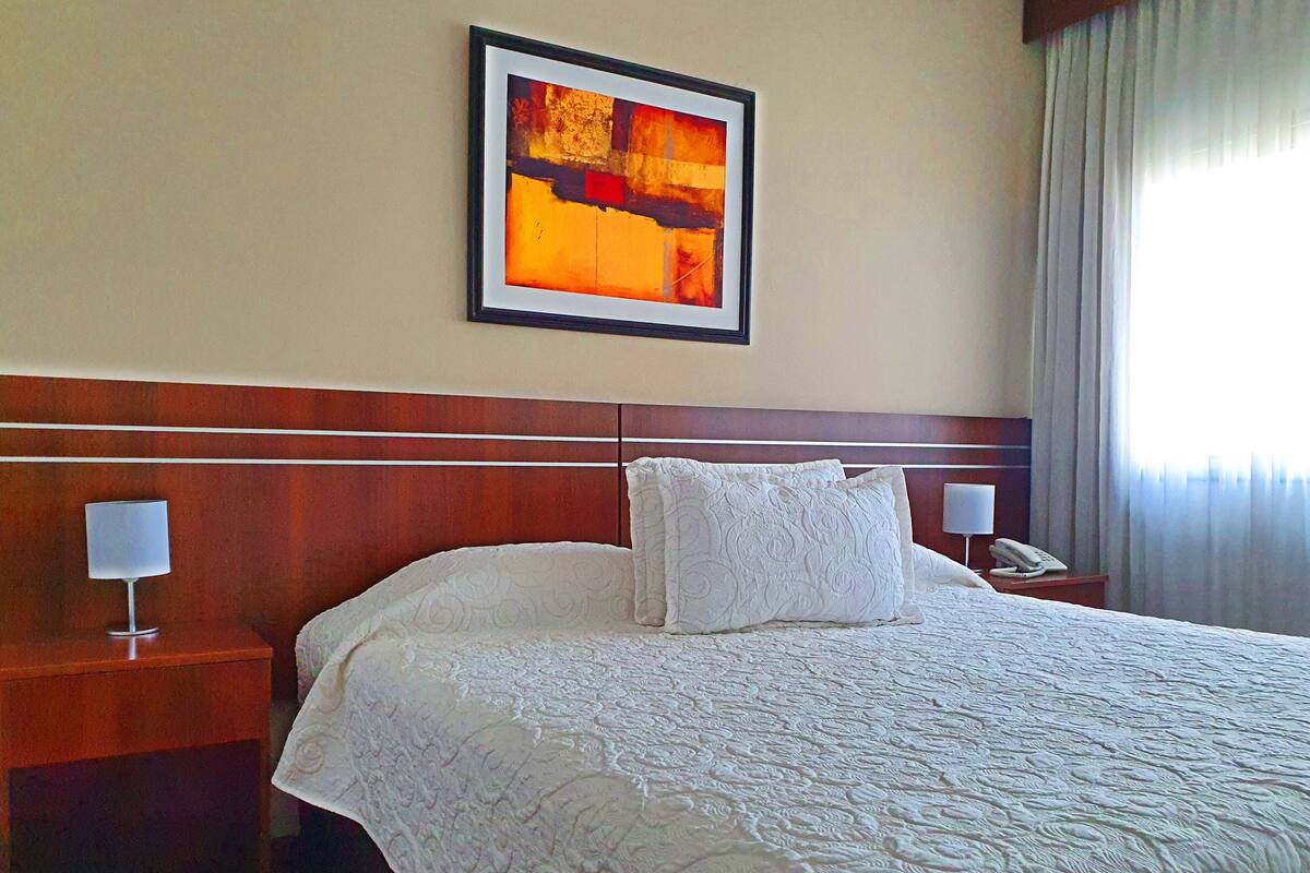 Habitación de hotel con una cama grande, una obra de arte enmarcada en la pared, una lámpara de noche y un teléfono en la mesita.