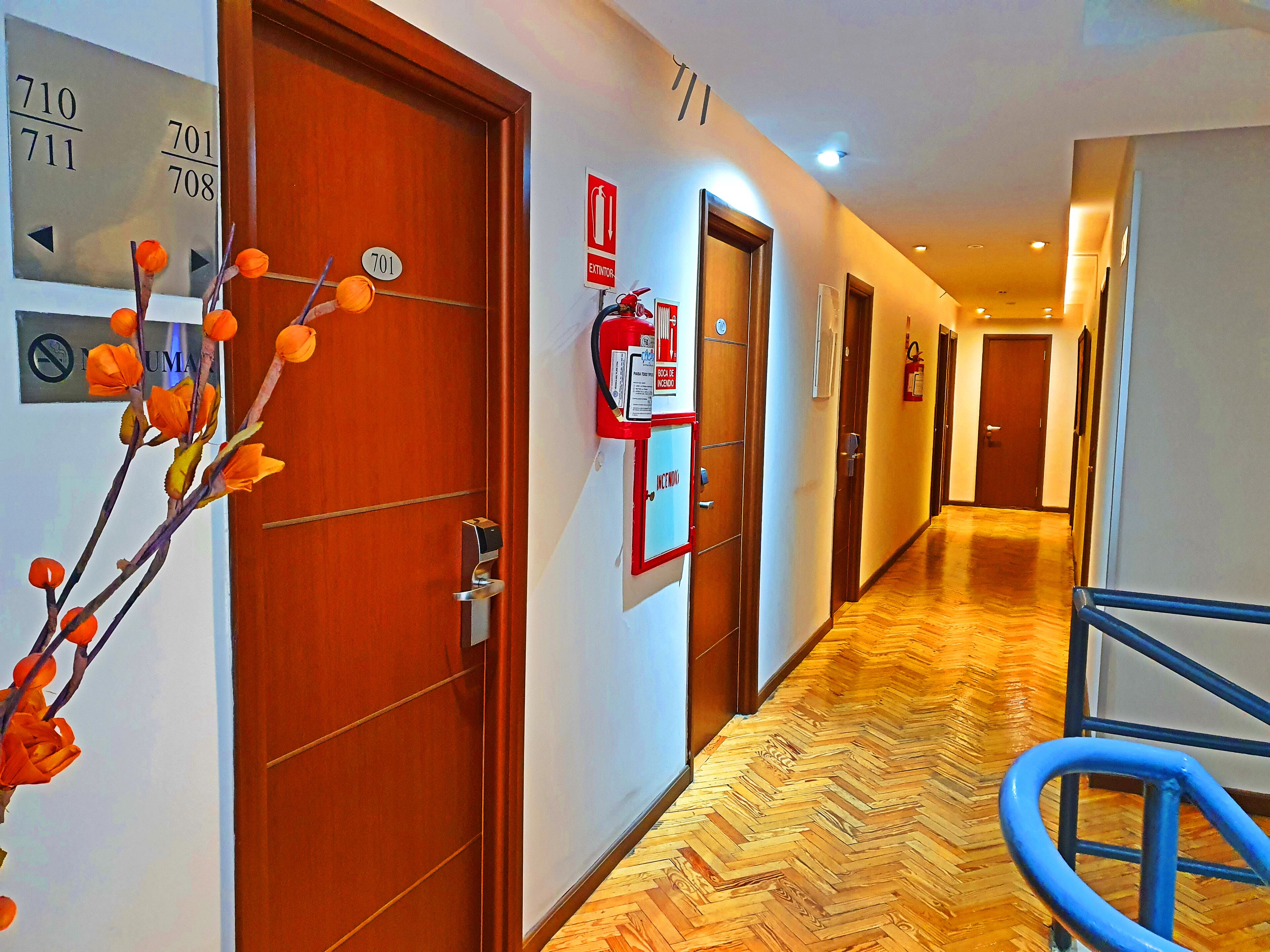 Pasillo de hotel con pisos de madera, puertas numeradas, un extintor y una barandilla azul.