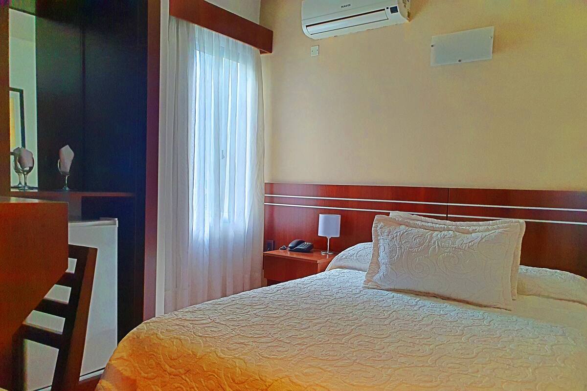 Habitación de hotel con cama, escritorio, silla, mesita de noche, lámpara y equipo de aire acondicionado.