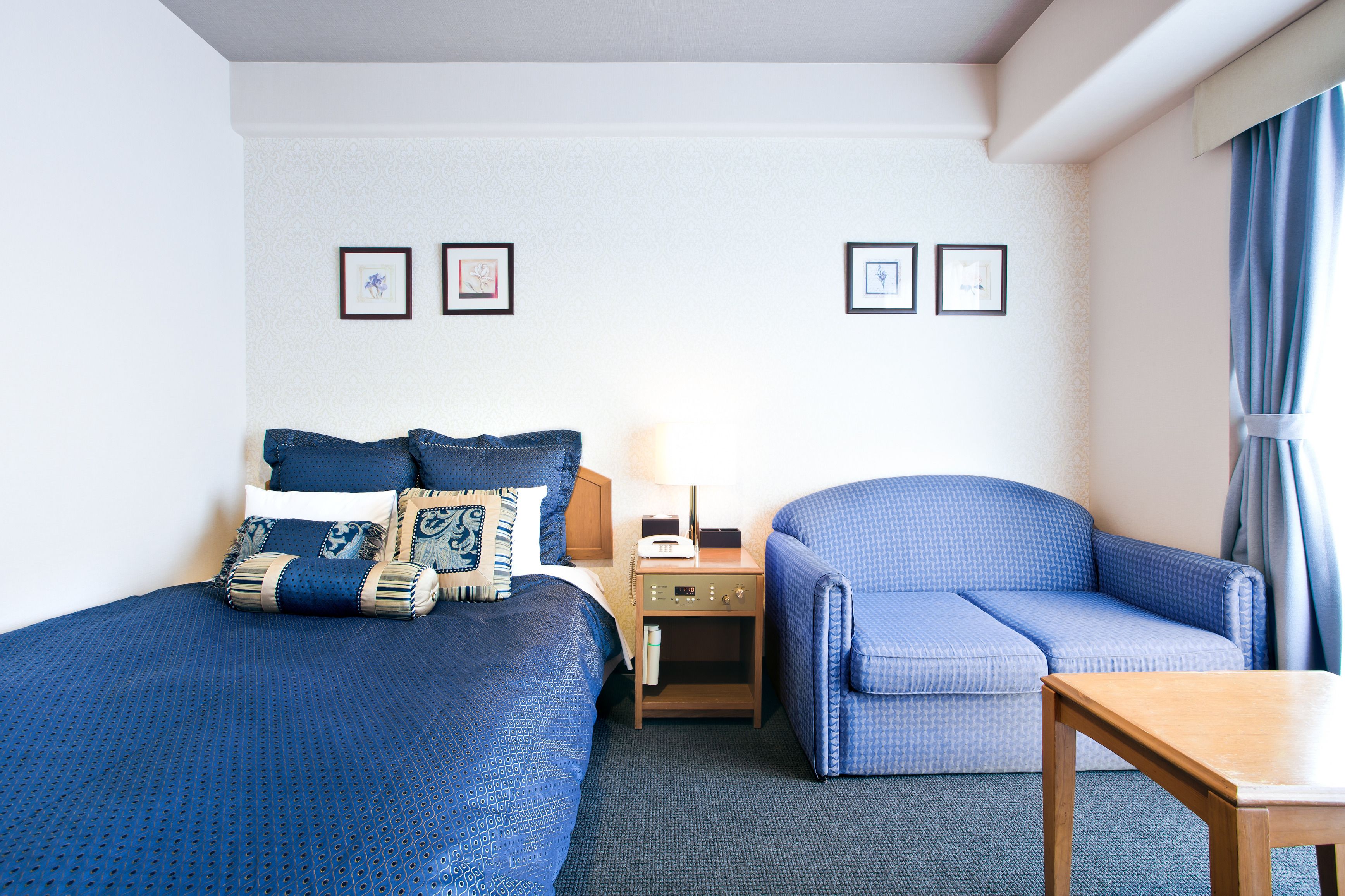 Quarto de hotel com uma cama, uma cadeira, uma mesa de cabeceira e uma mesa. Há duas quadros na parede.