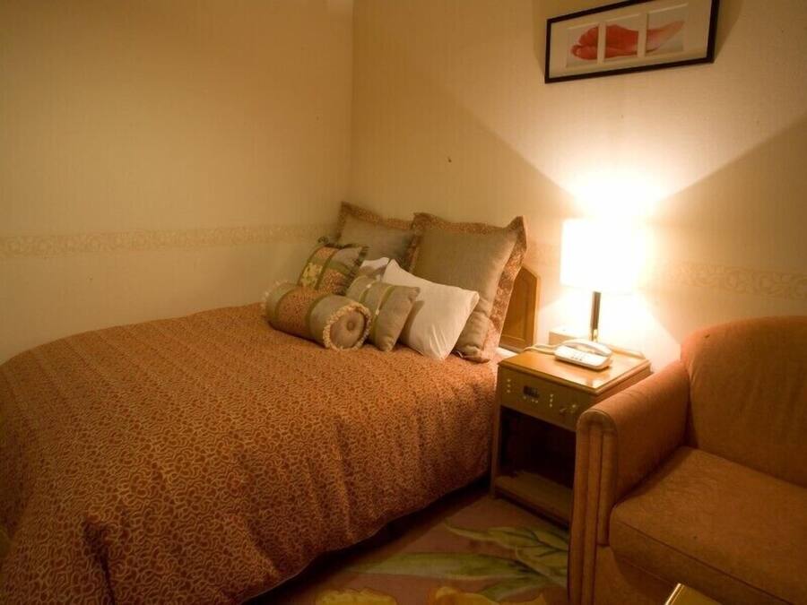 A single bed with a patterned bedspread, a nightstand with a lamp, and a chair.