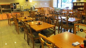 A spacious dining area with multiple wooden tables and chairs, a kitchen area in the background, and large windows providing natural light. - Ace Inn Kariya (Kariya)