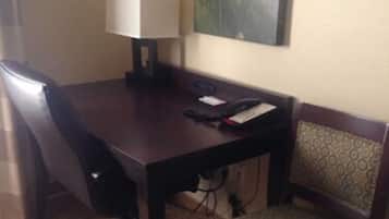 Habitación de hotel con un escritorio de madera, una silla, una lámpara, un teléfono y un cuadro de un paisaje montañoso.