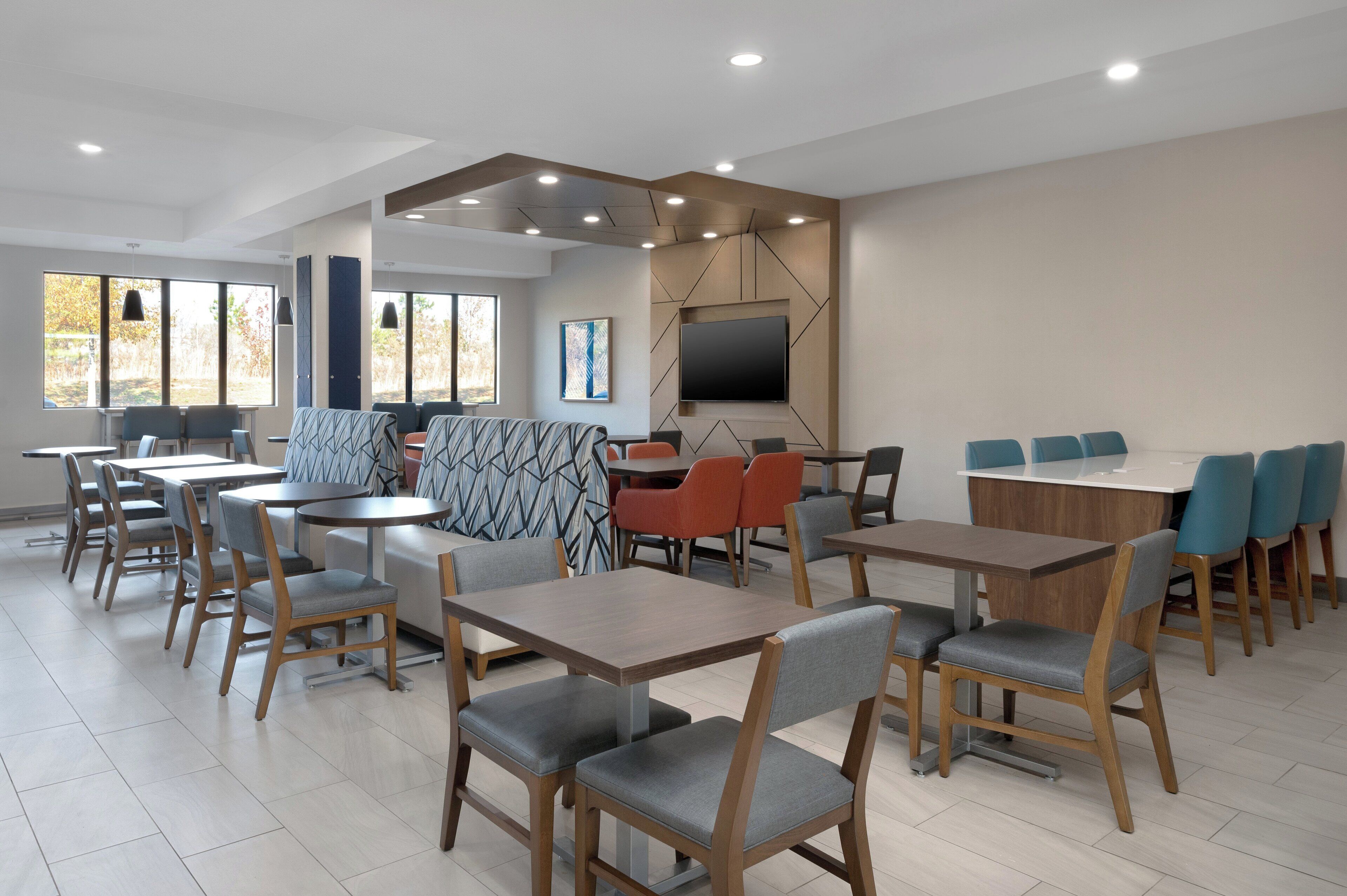 A modern dining area with multiple tables and chairs, a large flat-screen TV mounted on the wall, and a view of the outdoors through large windows.