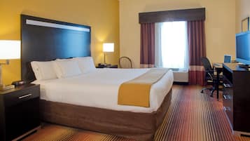 A hotel room with a large bed, a desk with a computer, a chair, a lamp, and a window with curtains.