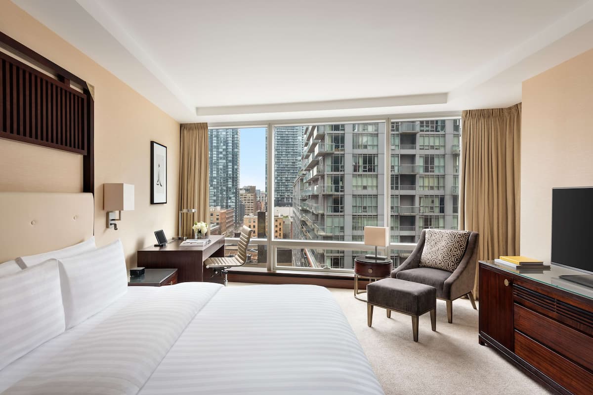 A hotel room with a large bed, a desk, a chair, a TV, and a view of tall buildings.