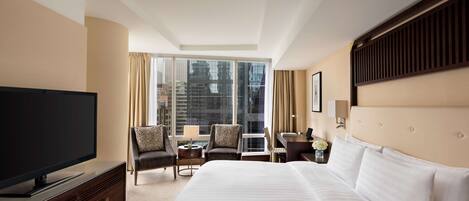 Ein Hotelzimmer mit einem großen Bett, einem Flachbildfernseher, einem Schreibtisch mit Stuhl und Blick auf die Stadt durch große Fenster.