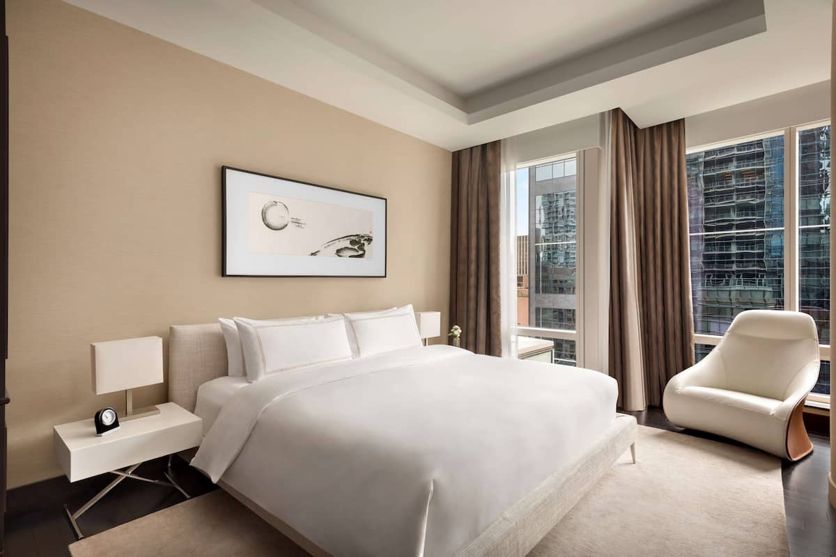 A modern hotel room with a large bed, a nightstand with a lamp, a chair, a framed picture on the wall, and a large window with curtains.