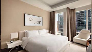 A modern hotel room with a large bed, a nightstand with a lamp, a chair, a framed picture on the wall, and a large window with curtains.