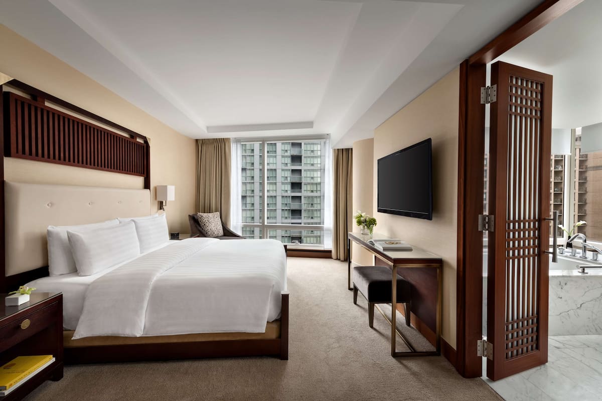 A hotel room with a large bed, a desk, a TV, and a view of high-rise buildings.