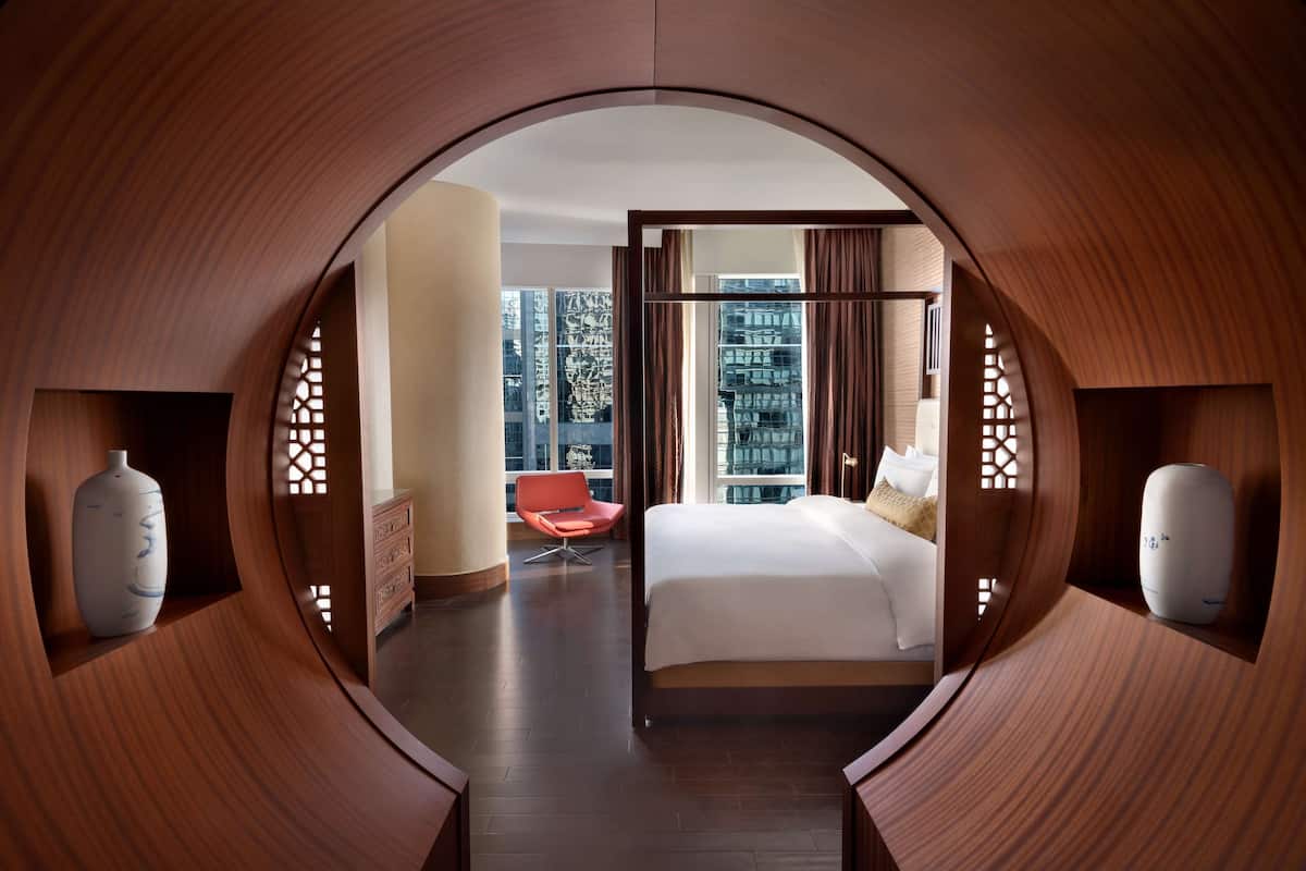 A modern hotel room with a curved wooden design, a bed, a red chair, and a view of the cityscape.