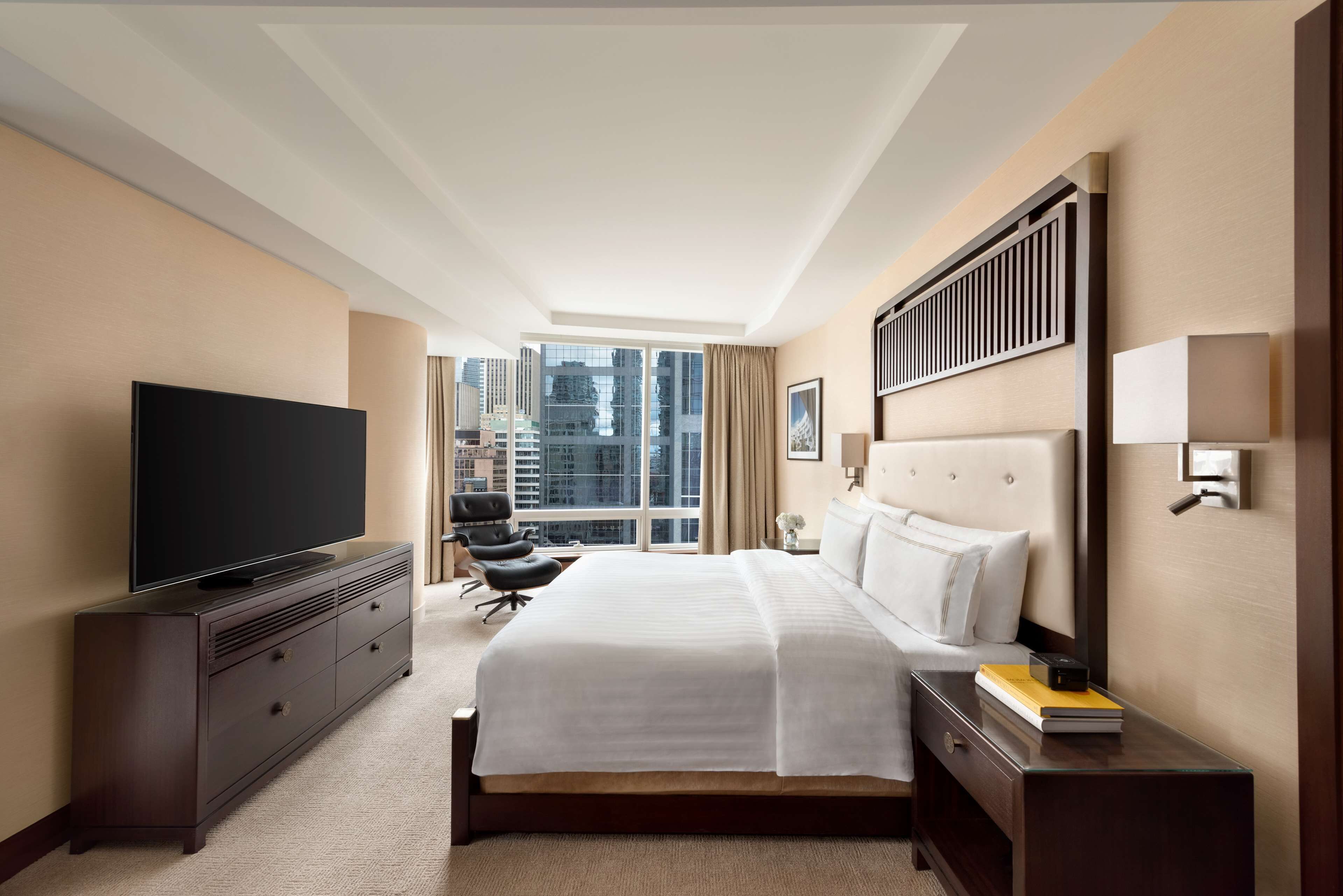 Ein Hotelzimmer mit einem großen Bett, einem Flachbildfernseher, einem Schreibtisch mit Lampe und Blick auf die Stadt durch große Fenster.