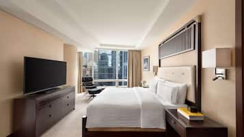 Ein Hotelzimmer mit einem großen Bett, einem Flachbildfernseher, einem Schreibtisch mit Lampe und Blick auf die Stadt durch große Fenster.