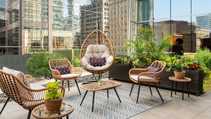 A rooftop terrace with wicker furniture, potted plants, and a view of city buildings.