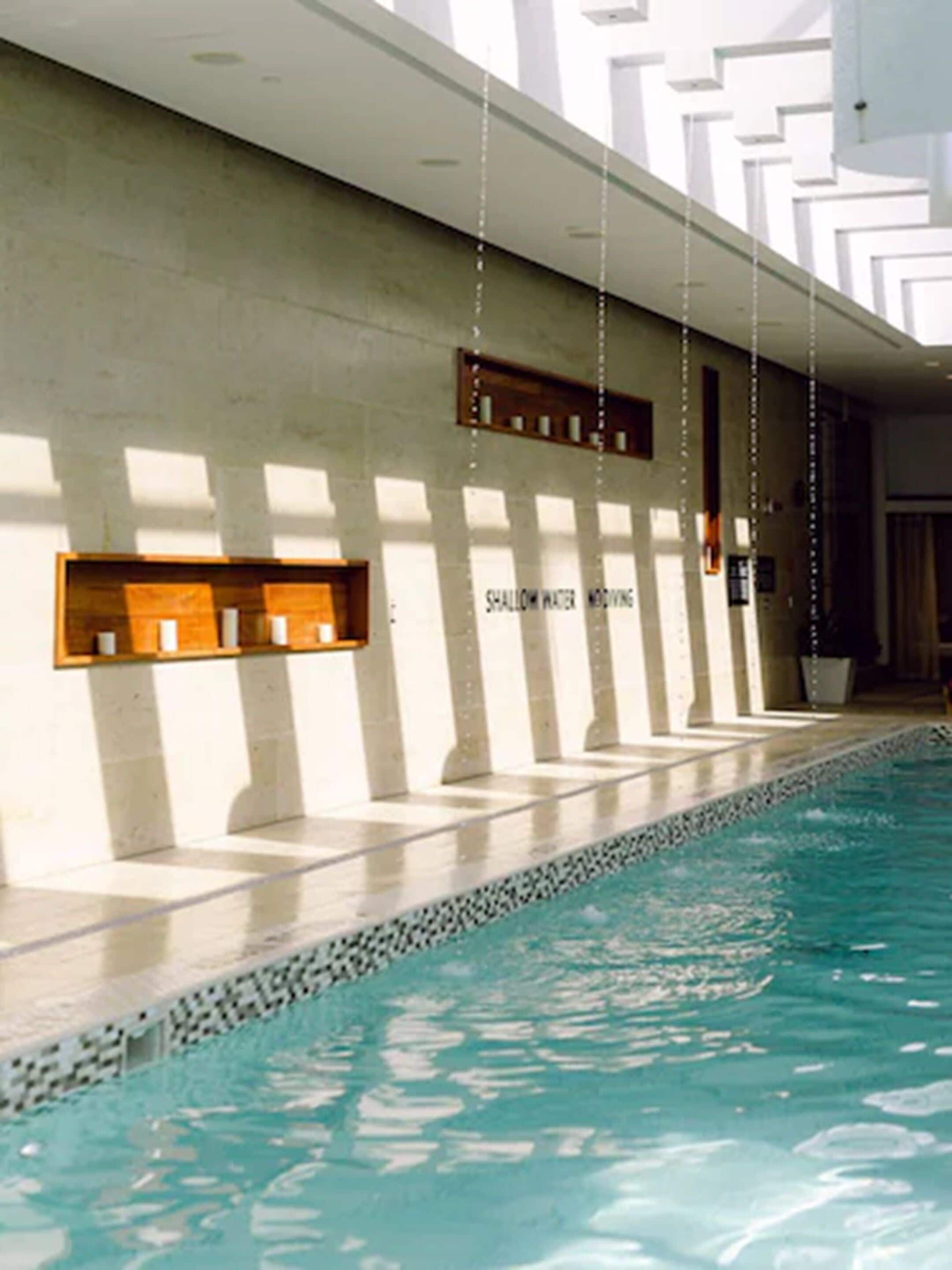 A swimming pool with a tiled edge and a concrete wall. There are wooden shelves on the wall and a sign that reads "SHALLOW WATER MEDUCTION".