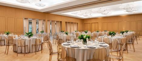 A large banquet hall with round tables, clear chairs, and chandeliers.