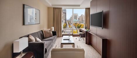 A modern hotel room with a large window, a sofa, a coffee table, a desk, and a TV mounted on the wall.