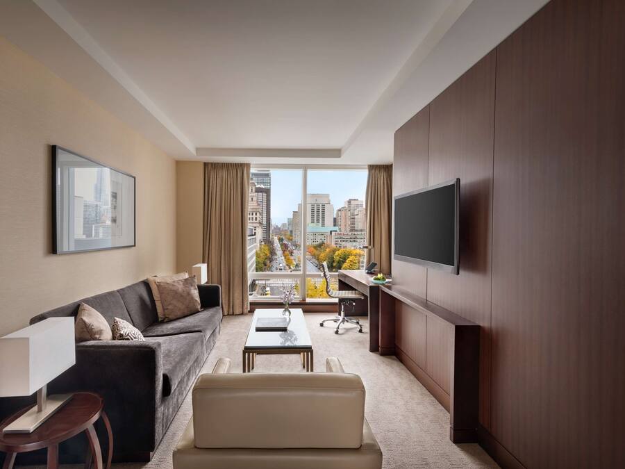 A modern hotel room with a large window, a sofa, a coffee table, a desk, and a TV mounted on the wall.