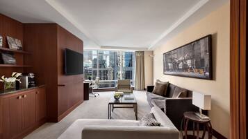 A modern hotel room with a large window offering a city view, a flat-screen TV mounted on the wall, a comfortable sofa, a small coffee table, and a wooden cabinet with a flat-screen TV.