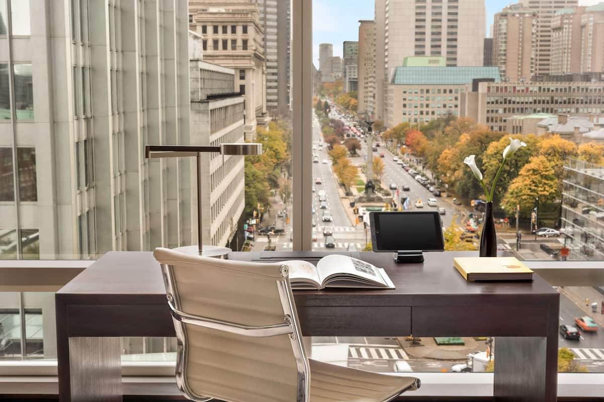 A modern office chair, a desk with a laptop, and an open book, with a cityscape view through a large window.